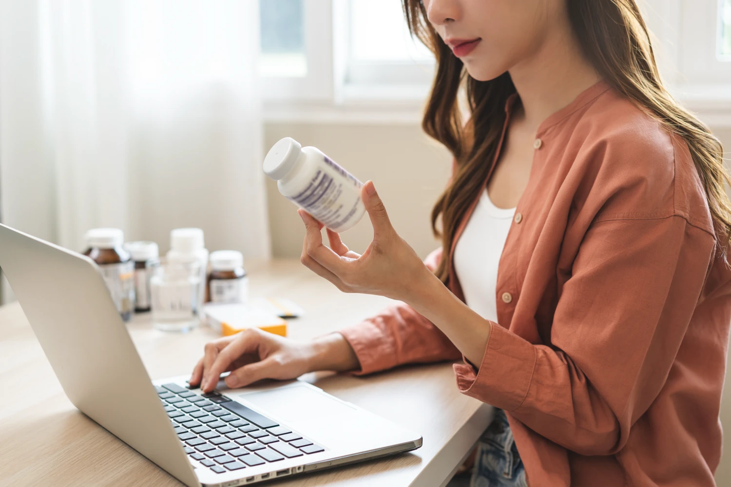 Woman sits at a laptop, holding a prescription bottle while looking at the screen.