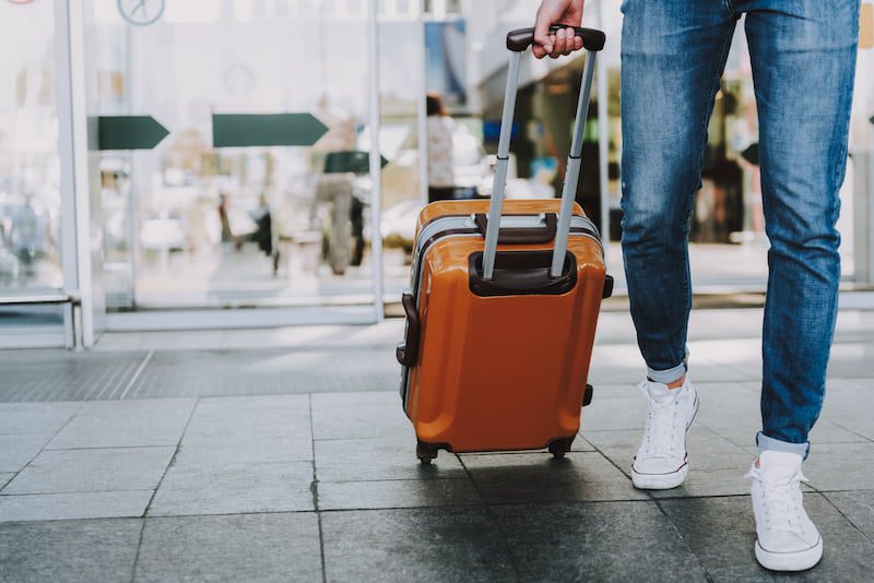 Close up of a man's shoes as he rolls his orange luggage out of an airport