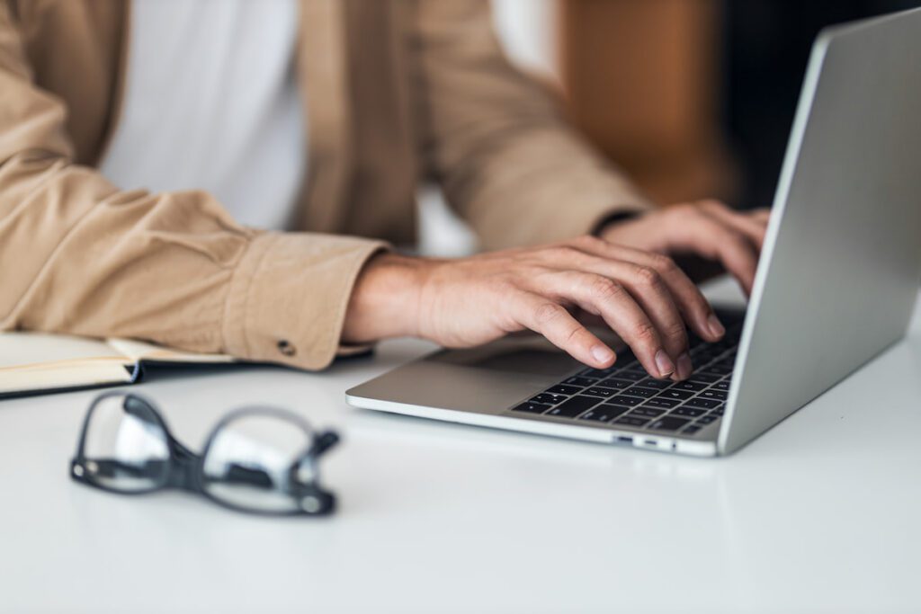 Person typing on a silver laptop at a white desk with glasses and an open notebook nearby.