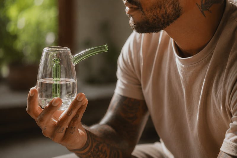 Man with tattooed forearms holding a glass water pipe filled with water in a sunlit room